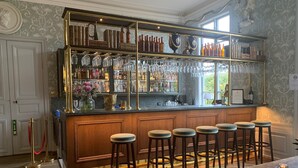 Un espace bar doté d’un comptoir en bois, de plusieurs tabourets et d’une vitrine en verre où sont exposées des bouteilles et des verres.