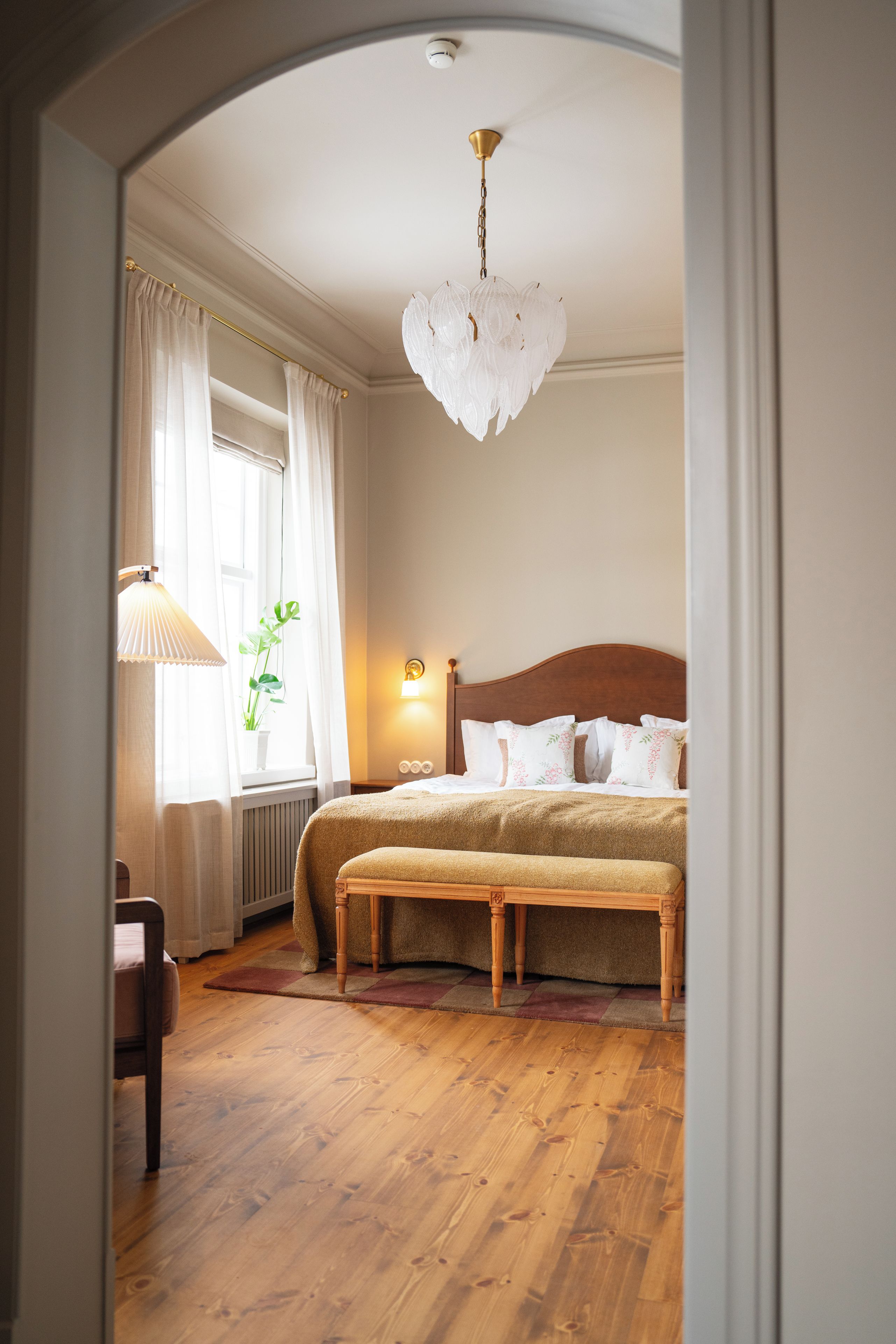 A bedroom with a wooden floor, a bed with a headboard, a bench, a lamp, and a window with curtains.