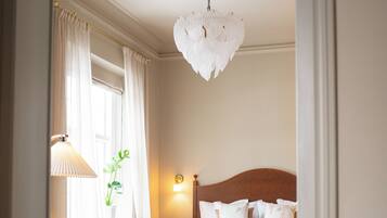 A bedroom with a wooden floor, a bed with a headboard, a bench, a lamp, and a window with curtains.