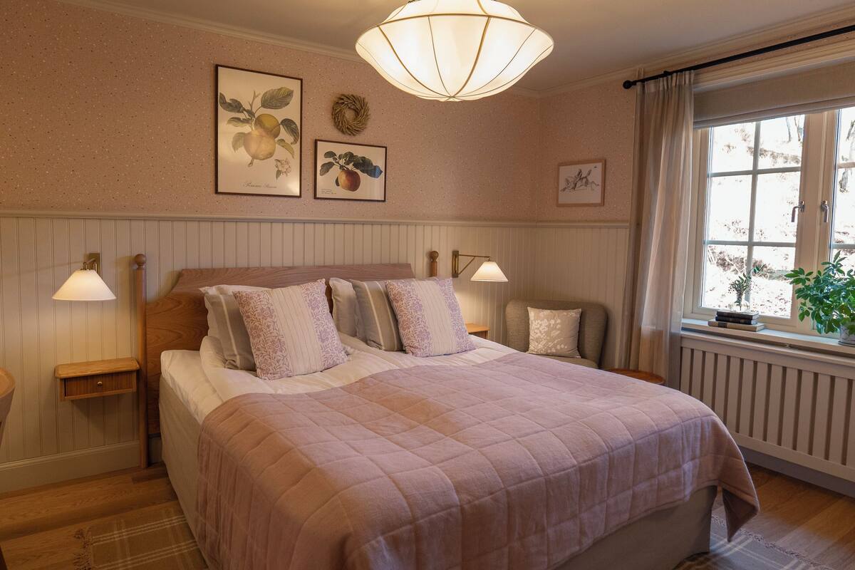 Une chambre à coucher avec un grand lit, deux tables de chevet, une lampe fixée au mur et une fenêtre donnant sur un beau paysage.