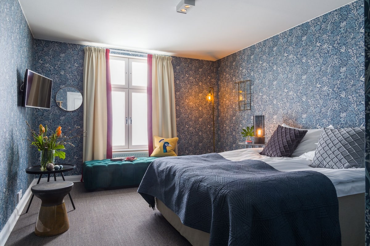 A bedroom with a bed, a bedside table, a mirror, a window with curtains, a wall-mounted TV, and a wall-mounted light fixture.