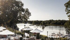 Un espacio al aire libre junto al lago con mobiliario de mimbre y sombrillas, con vistas a un lago tranquilo donde se pueden ver barcos amarrados.