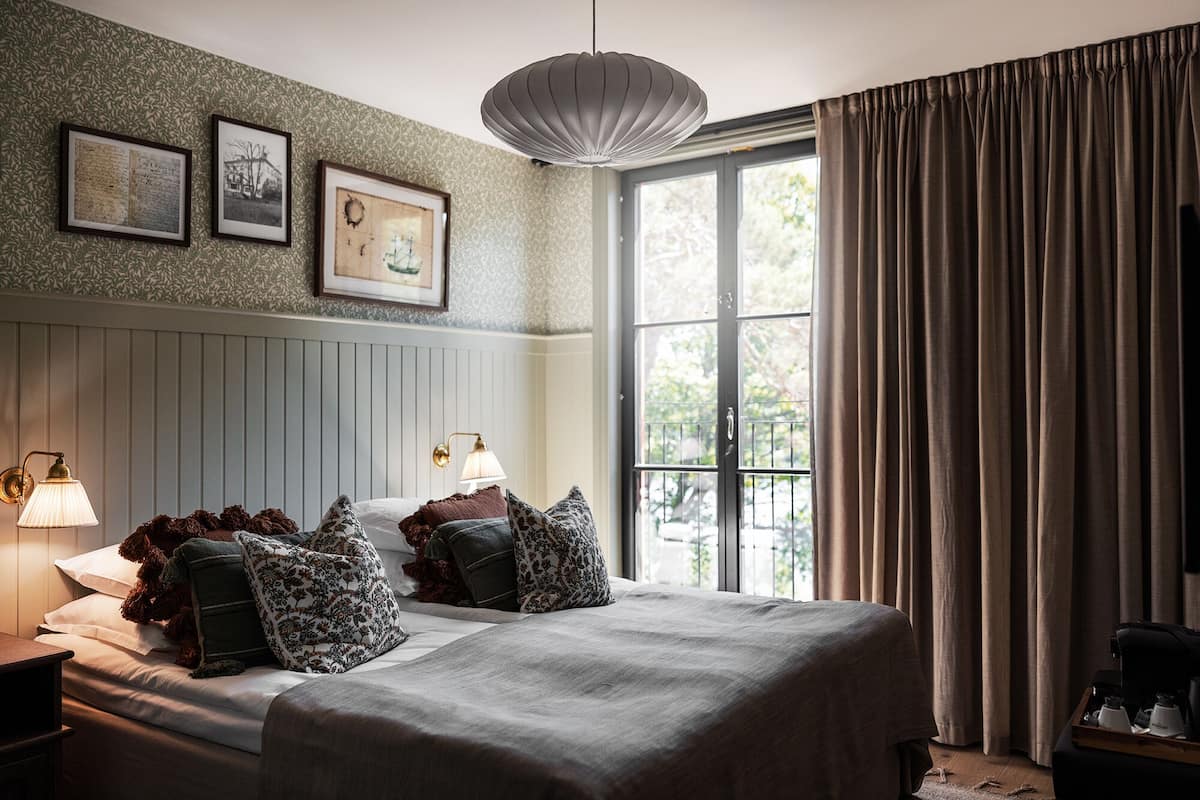 A bedroom with a bed, two bedside lamps, a window with curtains, and framed pictures on the wall.
