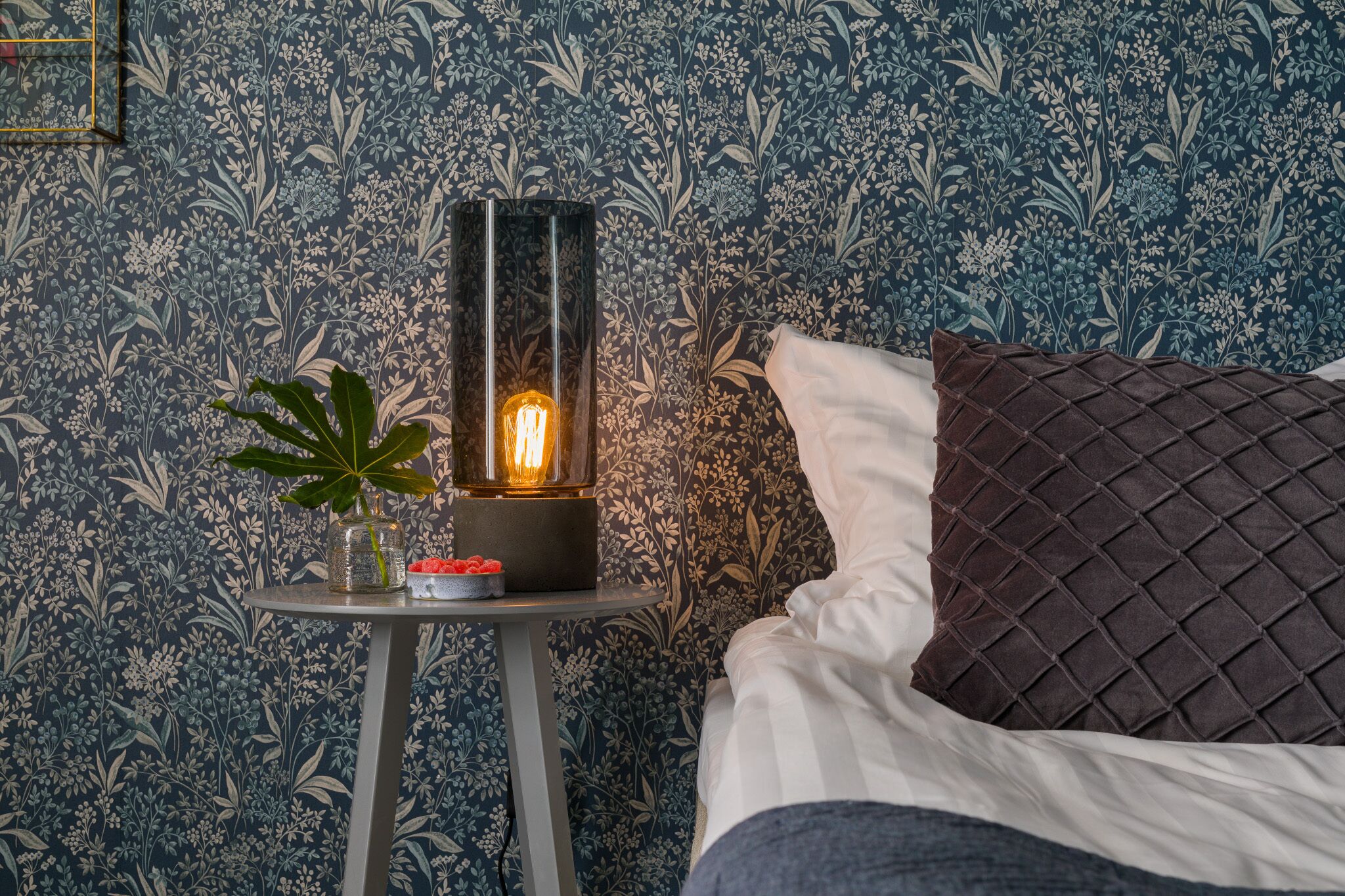 A room with a bed, a nightstand with a lamp, and a floral wallpaper.