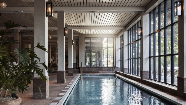 Una piscina interior con paredes de vidrio transparente, suelo de baldosas y techo de madera.