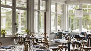 En restaurang med runda bord, trästolar och stora fönster med utsikt över grönområden.