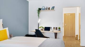 Une chambre d’hôtel moderne avec un lit, un bureau et une chaise. On trouve sur le bureau une petite plante, une horloge et une étagère avec des objets décoratifs.
