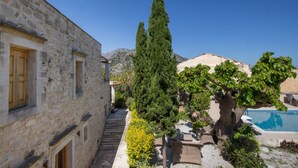 A stone building with wooden shutters, a tall coniferous tree, and a pool area with lounge chairs. - Arcus Suites (Rethymno)