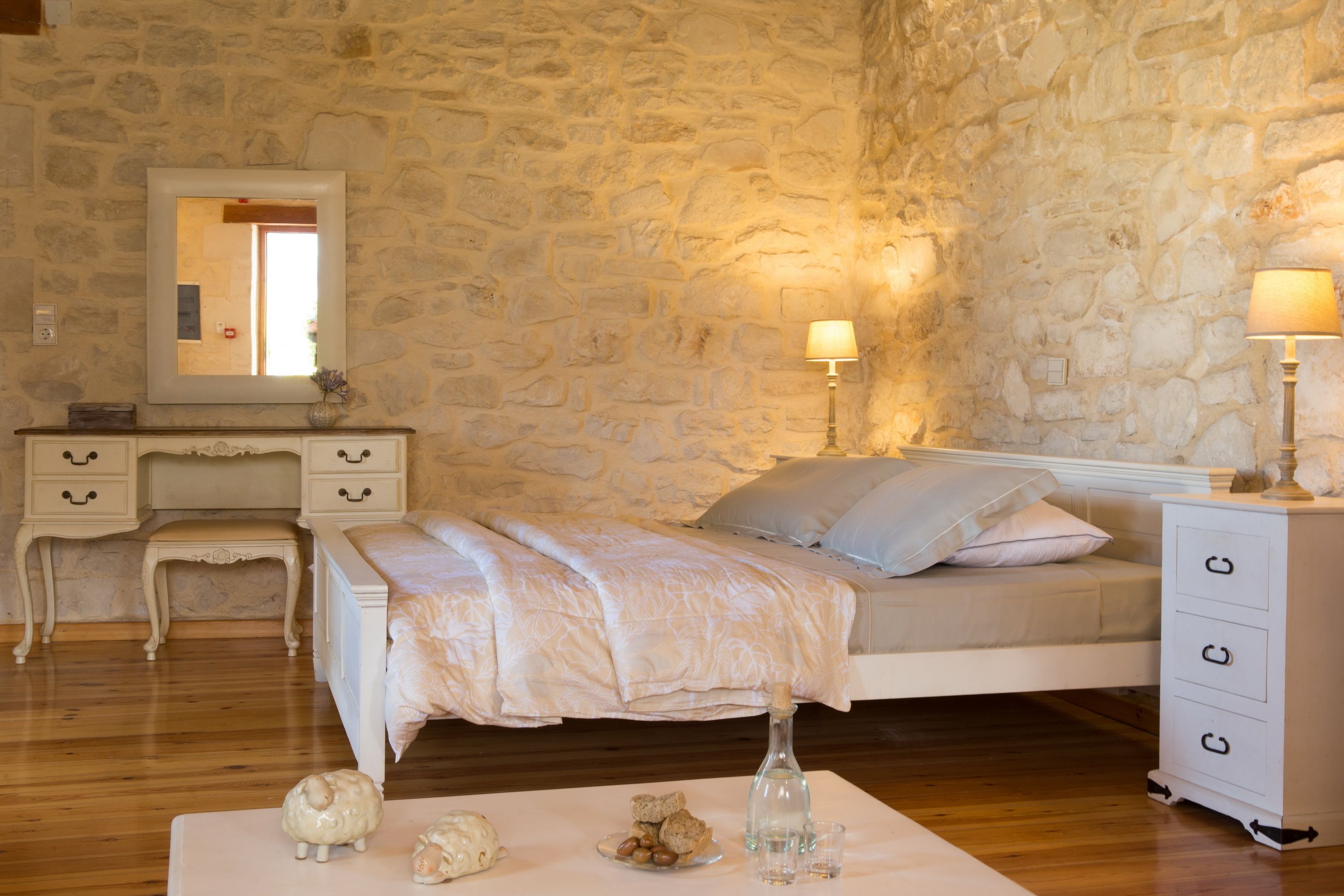 Een slaapkamer met een stenen muur, een bed met een wit hoofdeinde, een nachtkastje met een lamp, en een dressoir met lades.