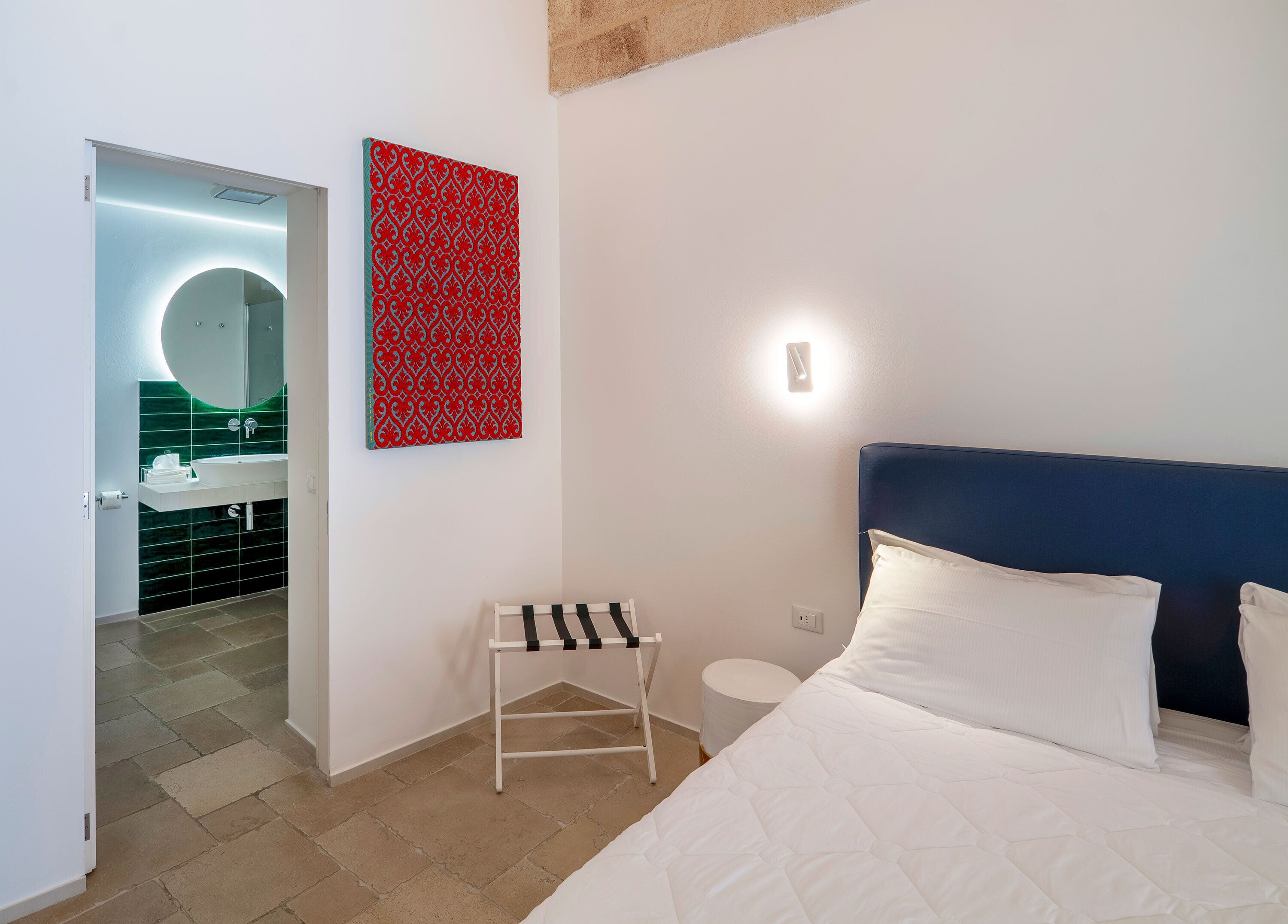 A modern hotel room with a bed, a bedside table, a small stool, a wall-mounted light, and a red patterned artwork.