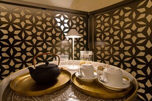 Une pièce dont un mur est décoré d’un motif géométrique, avec un plateau contenant un service à thé, une lampe blanche et une petite table sur laquelle sont posées des tasses et une théière.