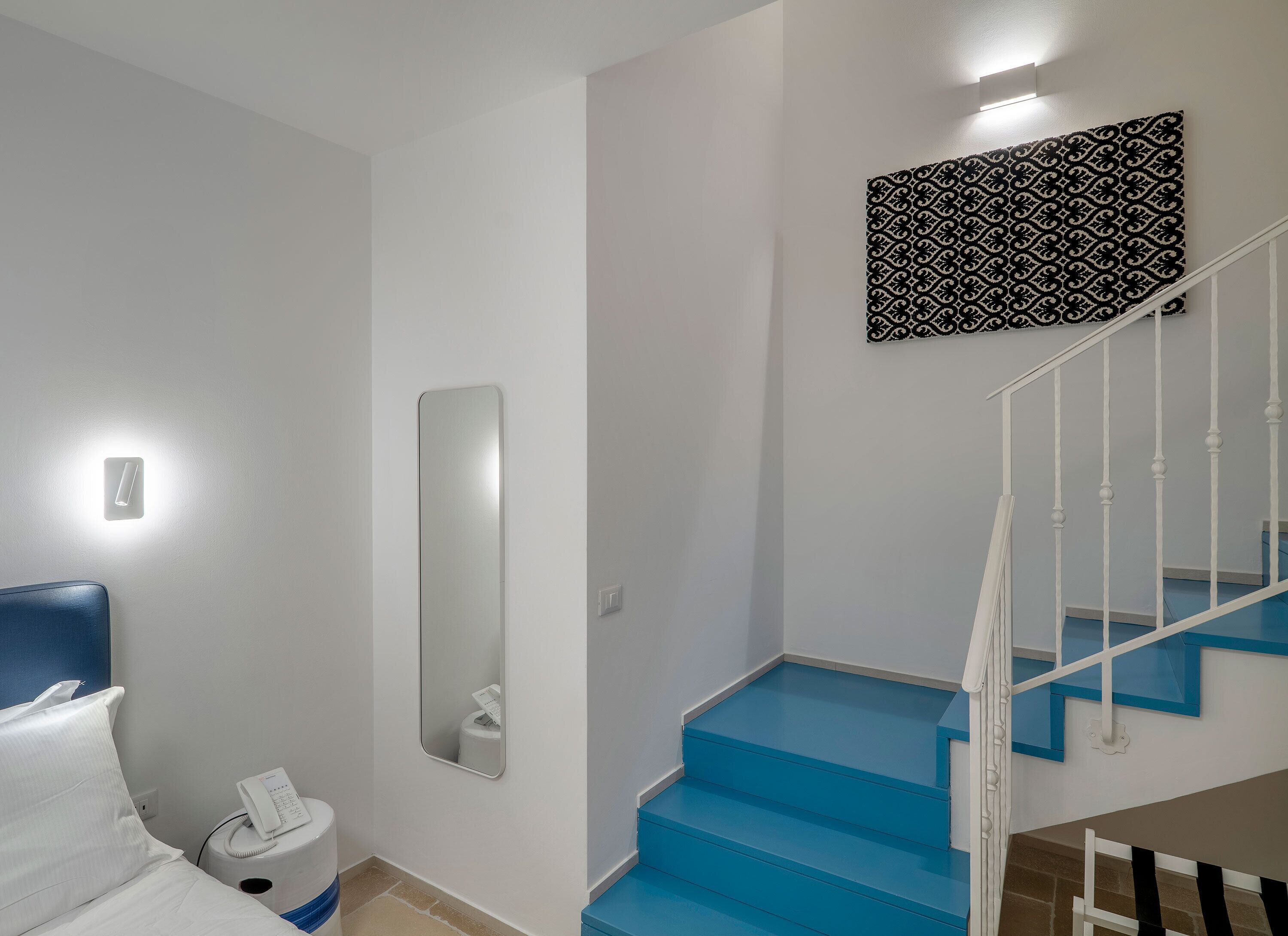 A hotel room with a bed, a bedside table with a phone, a mirror, a blue staircase, and a patterned wall decoration.