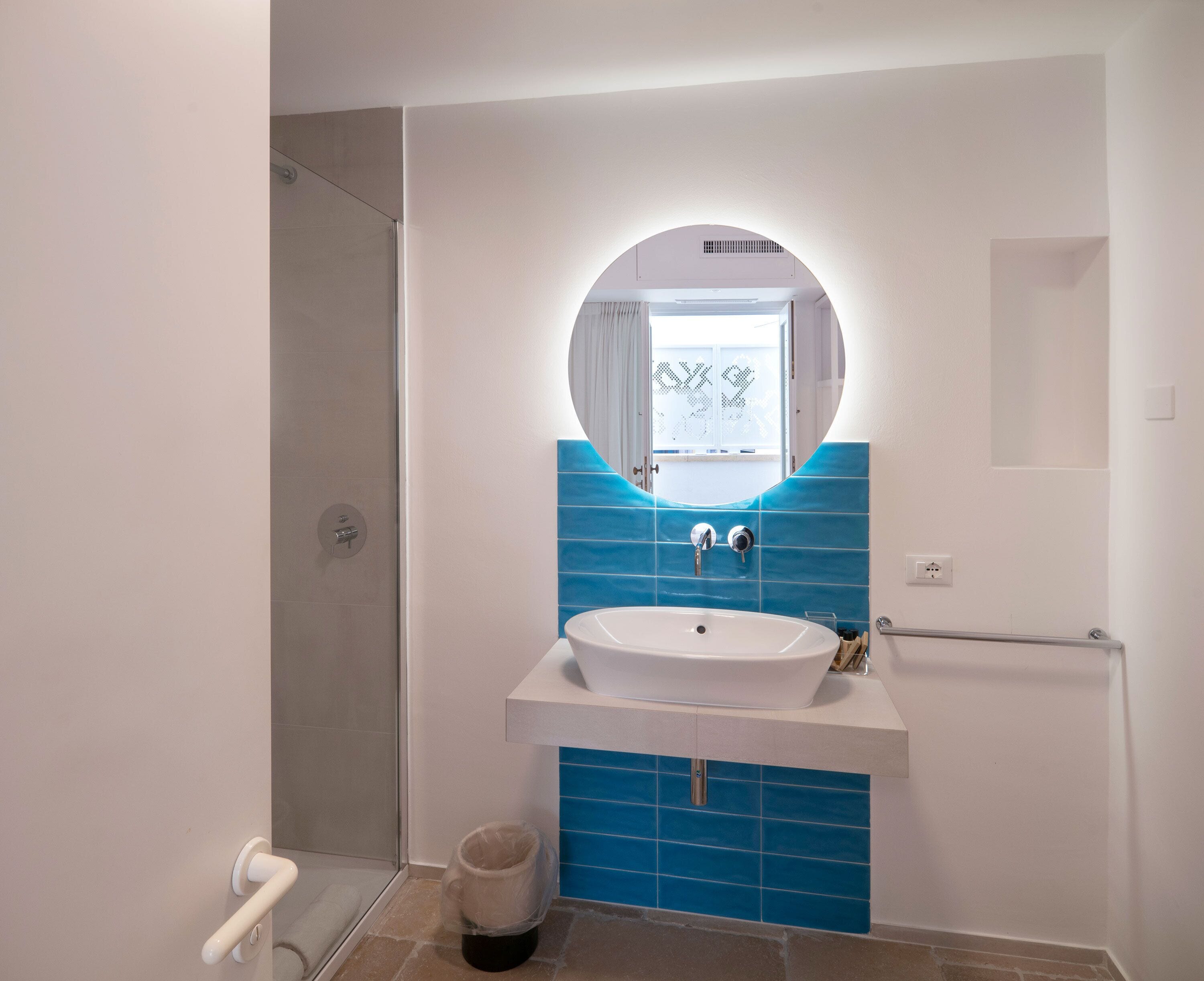 A bathroom with a white sink, blue tiles, a round mirror, and a glass shower enclosure.