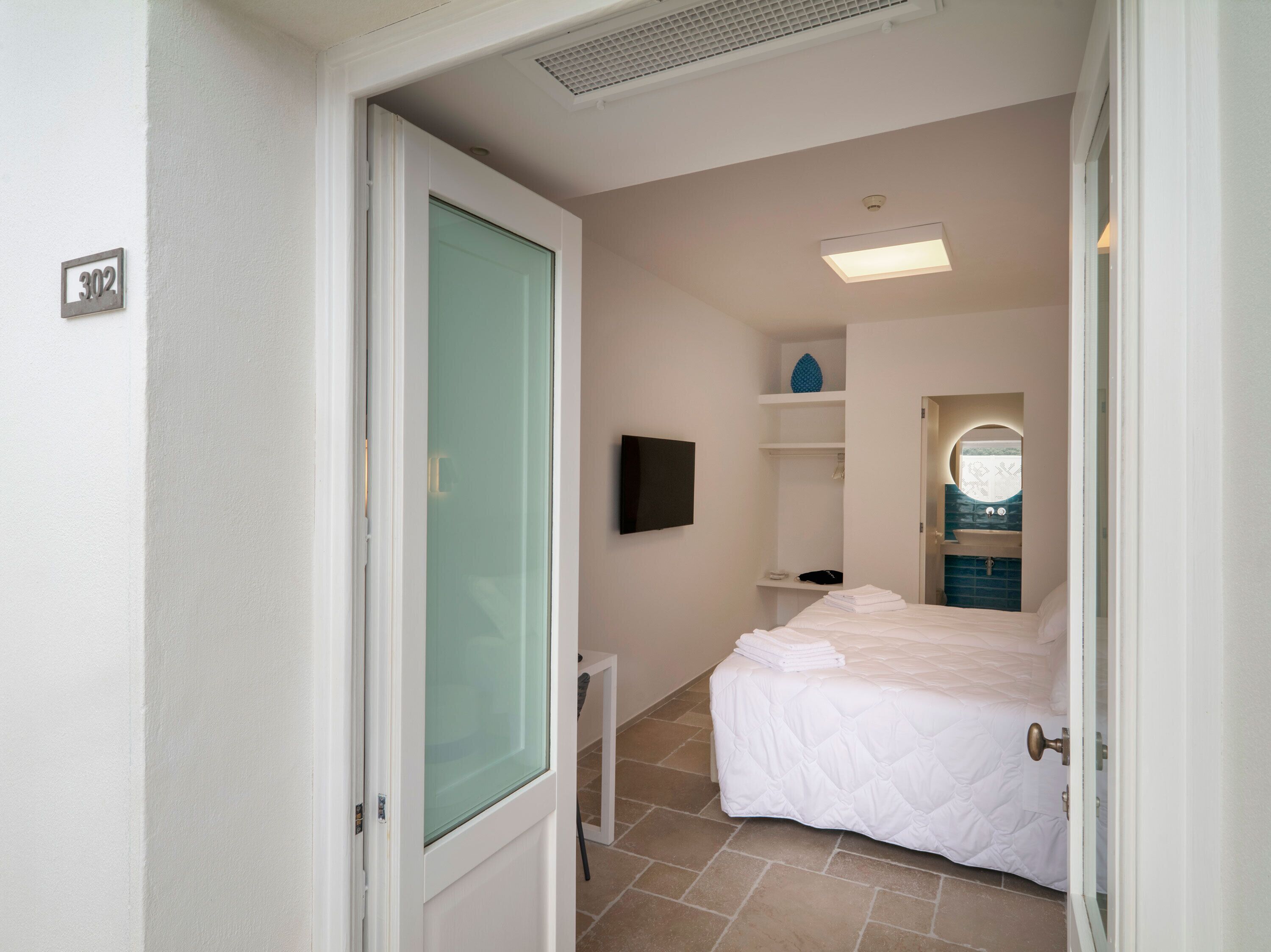 A hotel room with a bed, a television, a bathroom with a mirror, and a tiled floor.