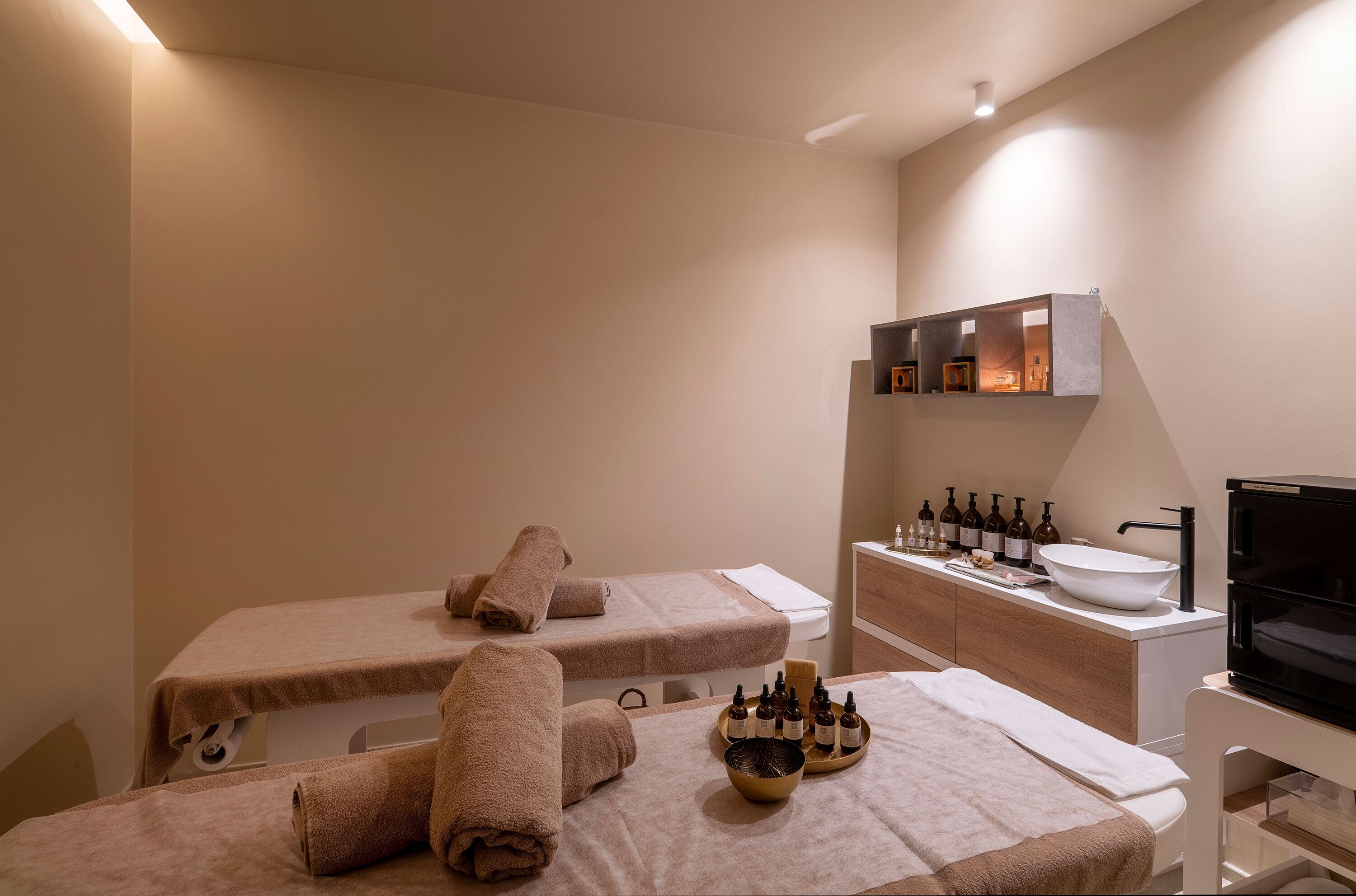 Un cuarto de spa con dos camillas de masaje, toallas, botellas de aceites y un lavamanos.