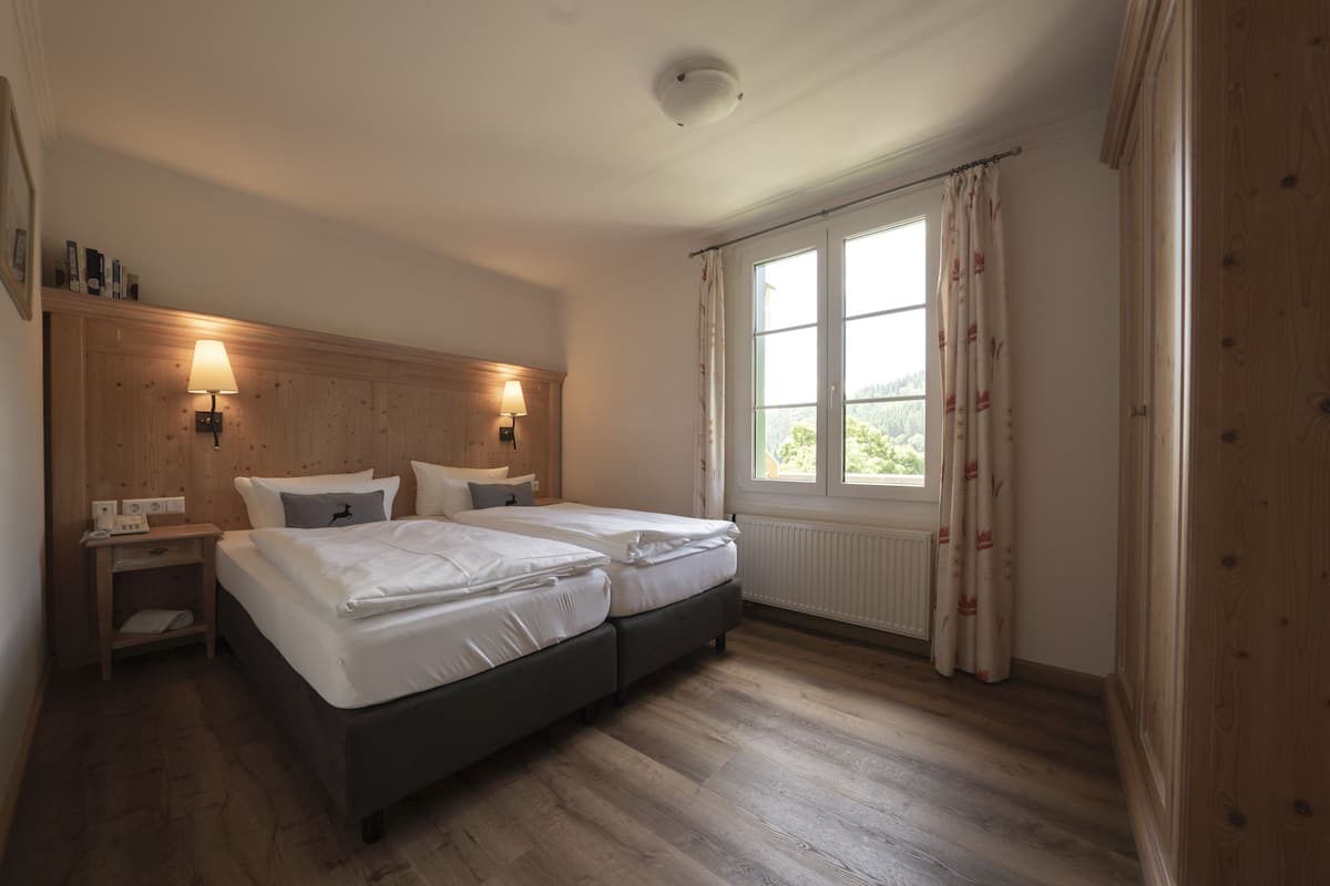 Un dormitorio con cabecera de madera, dos camas, lámparas de noche, una ventana con cortinas y un radiador.