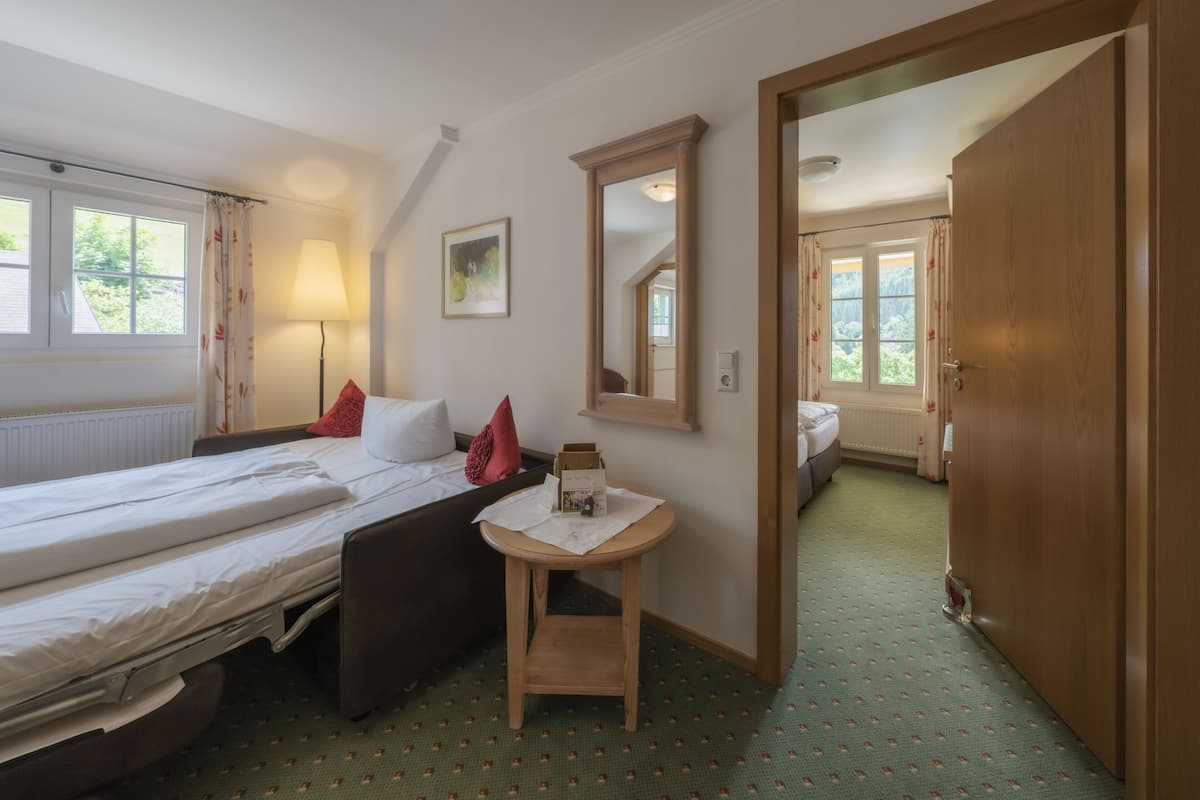 Habitación de hotel con dos camas, una mesita, un espejo y vista a otra habitación por una puerta abierta.