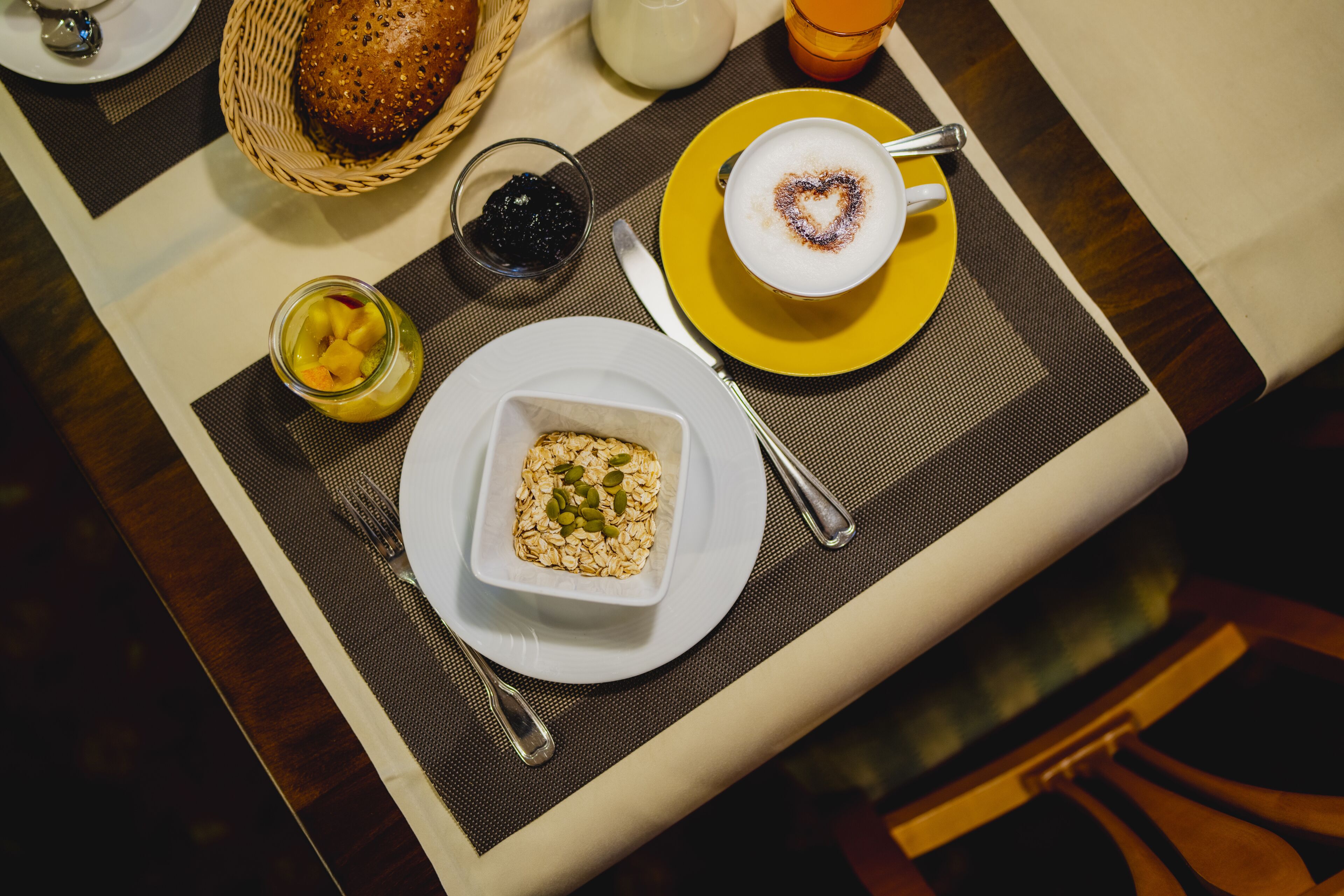 A breakfast setting with a basket of bread, a bowl of oatmeal, a glass of juice, and a cup of coffee with a heart-shaped design on the foam.
