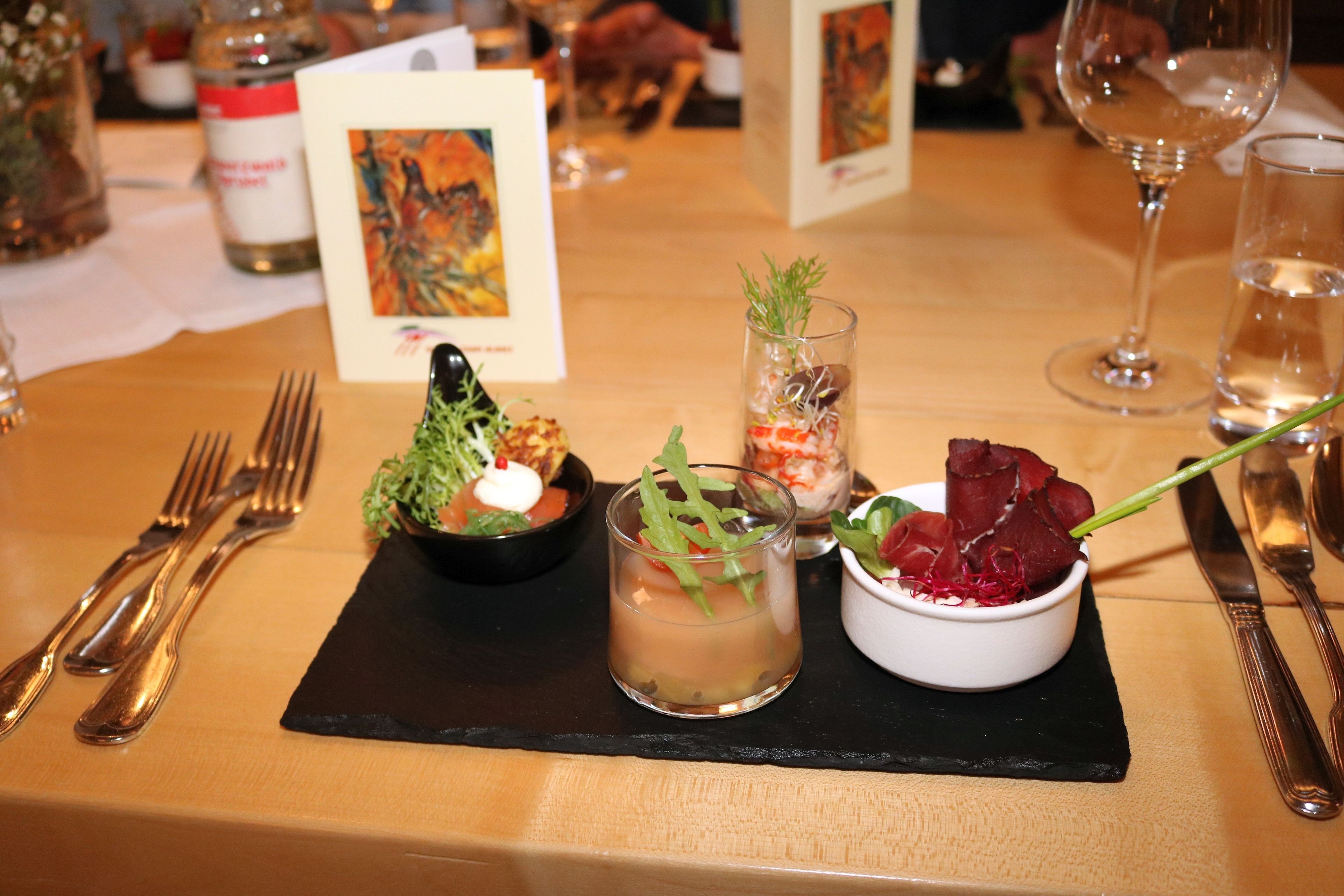 Een eettafel met een zwarte placemat, bestek, een glas water, een kleine schaal rode bessen en een glas met groene garnering.