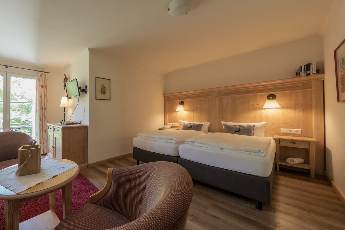 Habitación de hotel con dos camas, cabecero de madera, lámparas de noche, una mesita con lámpara y una silla.