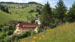 Ländliche Umgebung mit einem großen Gebäude, umgeben von Grünflächen, und einer hügeligen Landschaft im Hintergrund.