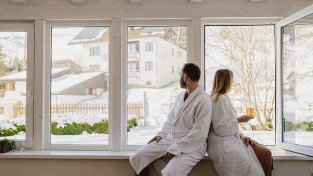 Zwei Personen in weißen Roben sitzen an einem Fenster mit Blick auf eine verschneite Landschaft.
