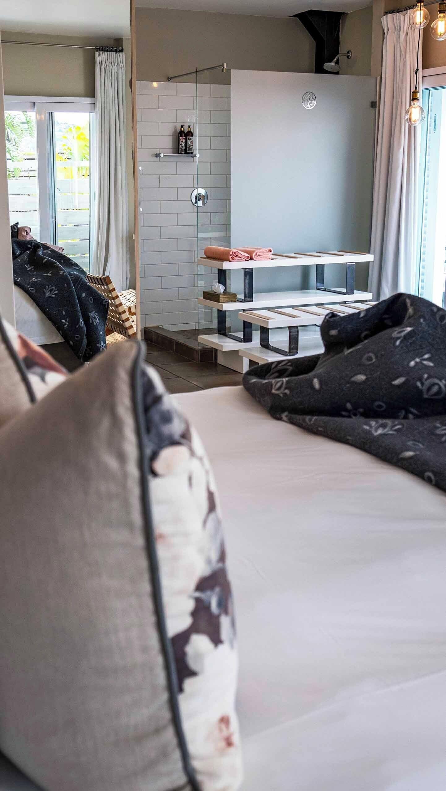 A bedroom with a bed, a bedside table, a window with curtains, and a shower area with a glass door.