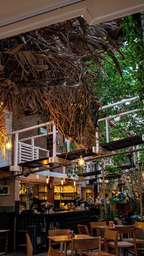 An indoor dining area with wooden tables and chairs, a bar with shelves of bottles, and hanging plants. - The Lofts Boutique Hotel (Knysna)
