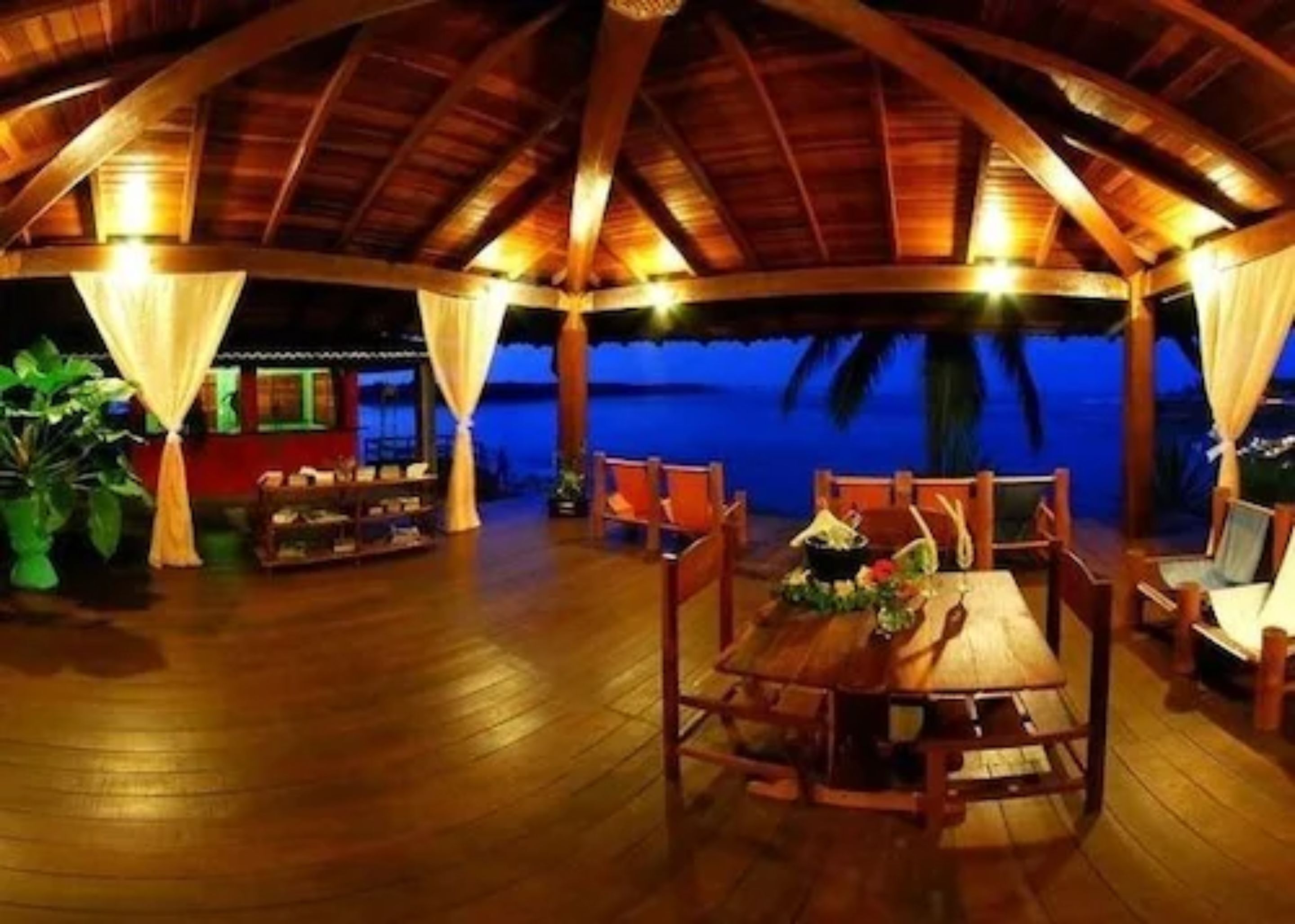 Een houten terras binnen met een tafel en stoelen, met uitzicht op een wateroppervlak bij nacht.