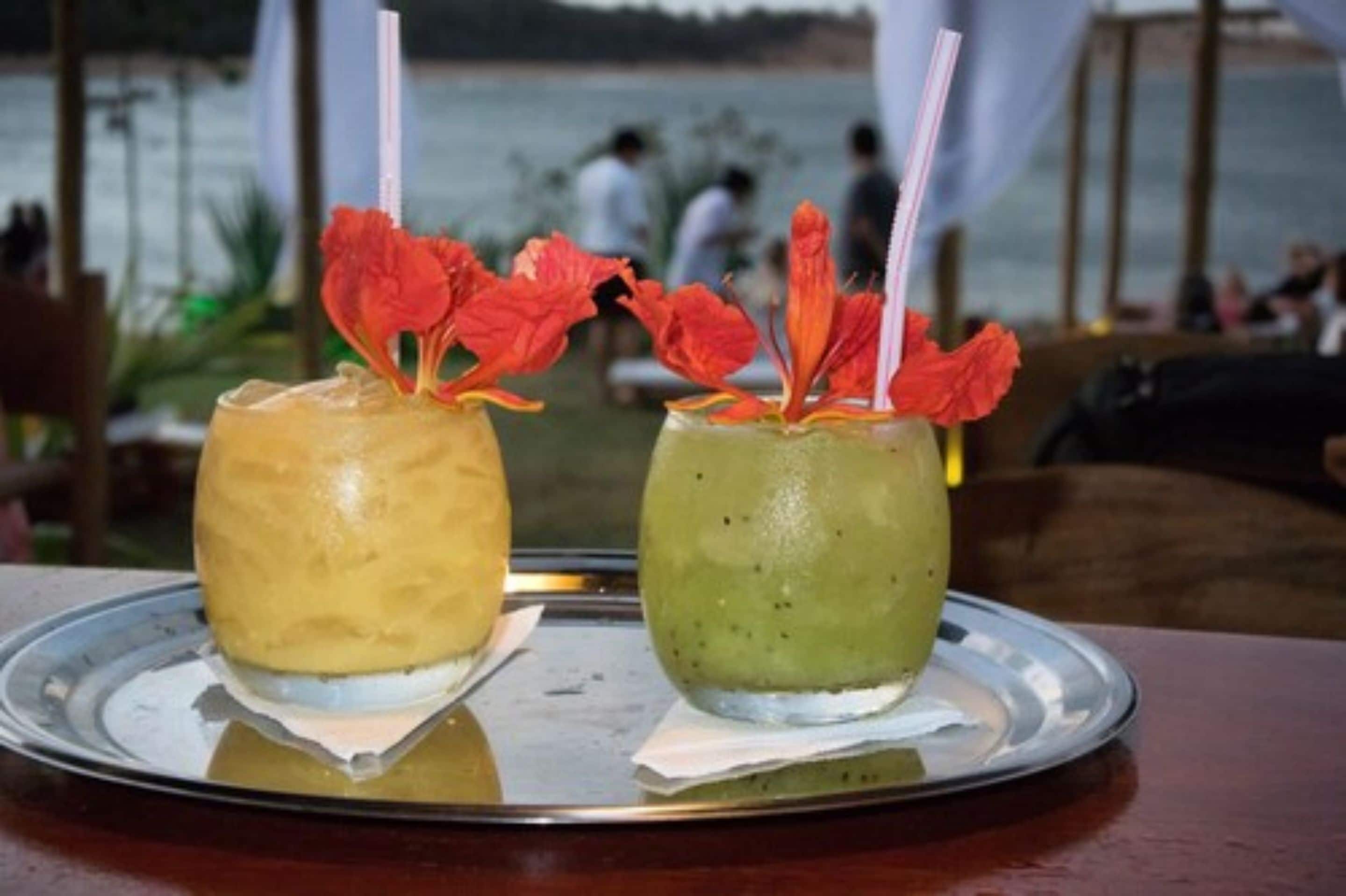 Dos bebidas sobre una bandeja, con vistas a un cuerpo de agua y personas al fondo.