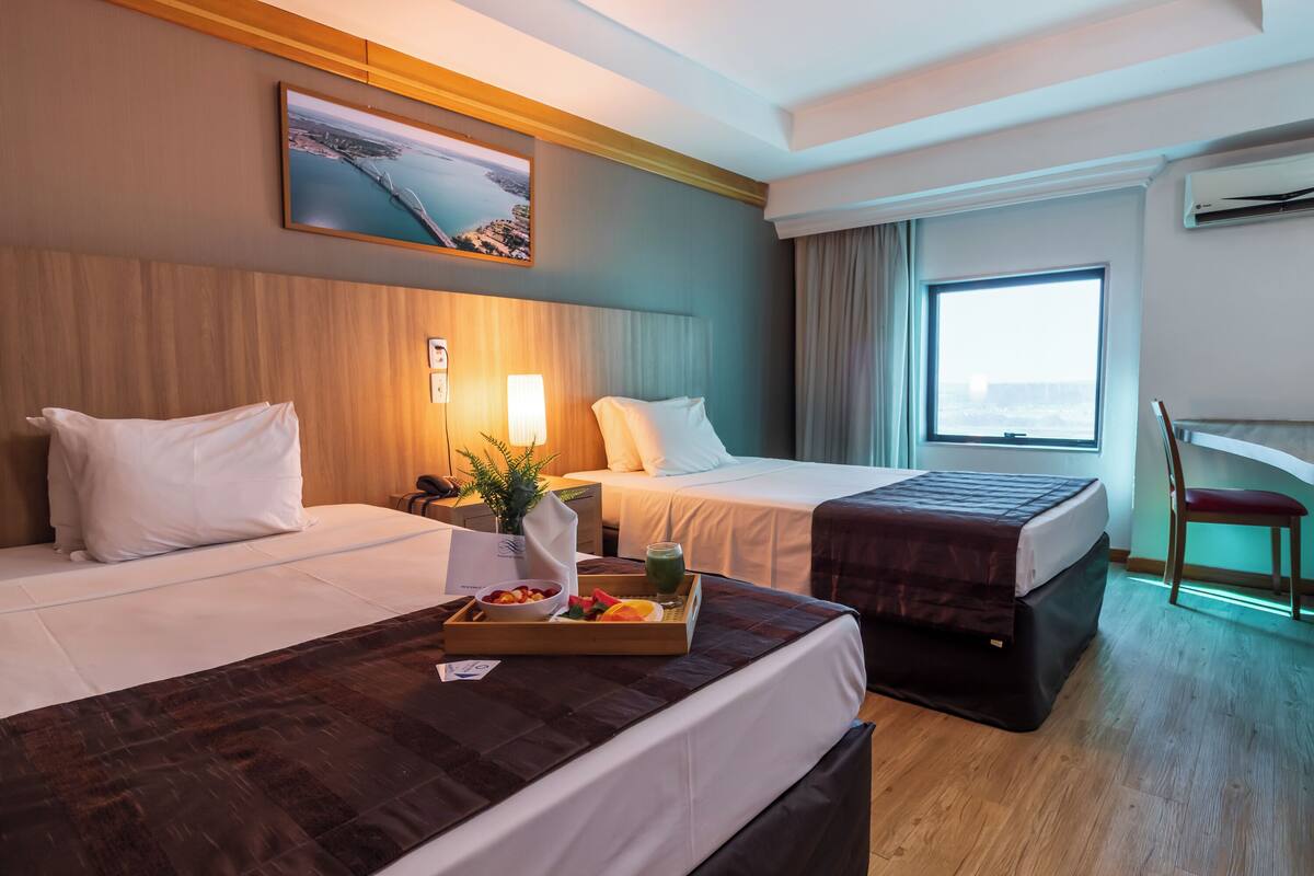 Ein Hotelzimmer mit zwei Betten, einem Schreibtisch, einem Sessel, einem Fenster mit Blick und einem Gemälde an der Wand.