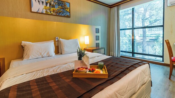 Habitación de hotel con una cama grande, una bandeja con frutas y una bebida verde, un cuadro en la pared y una ventana con vistas a árboles.