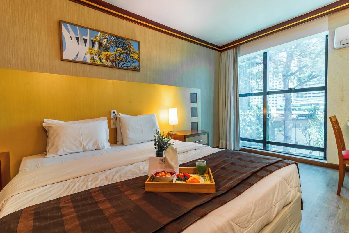 Ein Hotelzimmer mit einem großen Bett, einem Tablett mit Früchten und einem grünen Getränk, einem Gemälde an der Wand und einem Fenster mit Blick auf Bäume.