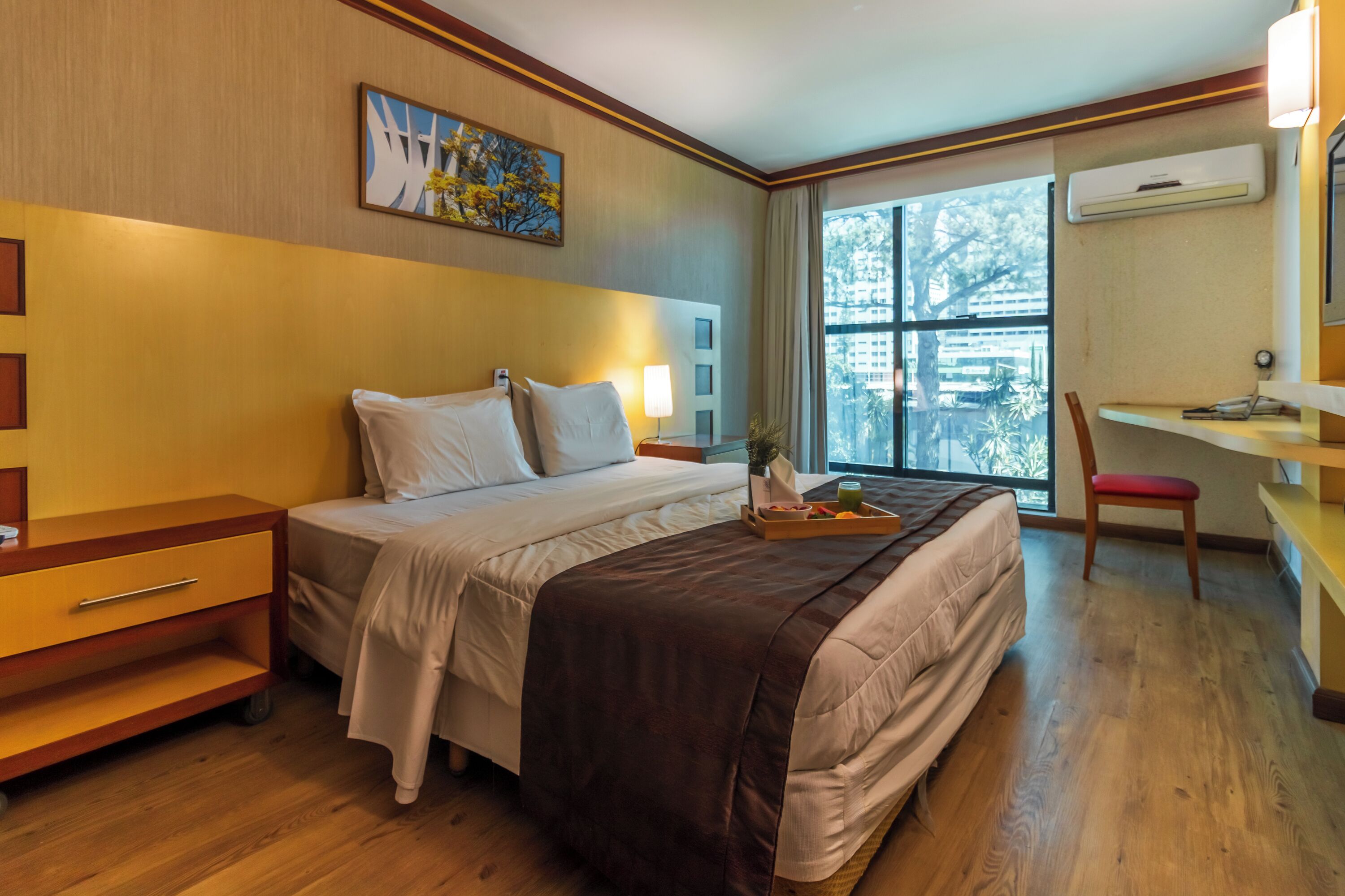 A hotel room with a bed, bedside table, lamp, window, and a desk with a chair.