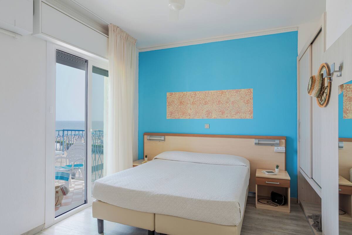 A hotel room with a bed, a bedside table, a nightstand, a window with curtains, and a view of the outside.