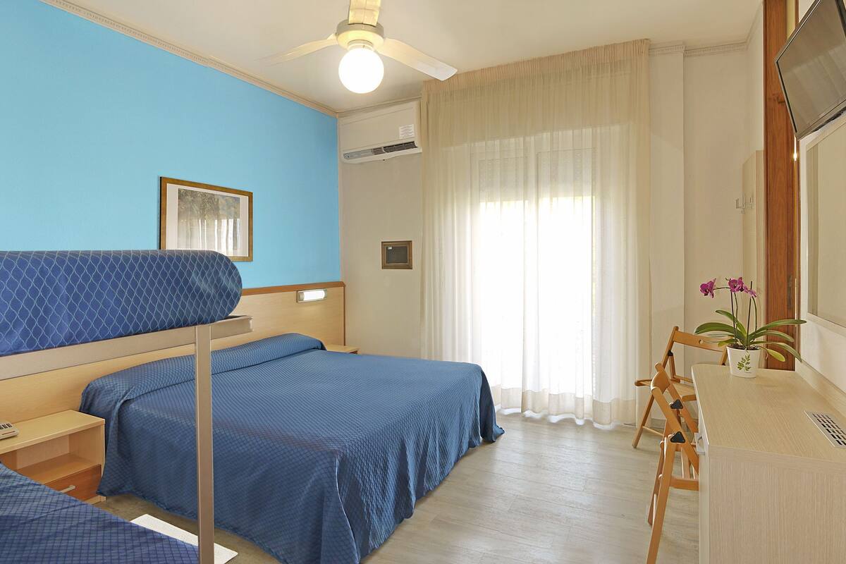 A hotel room with two beds, a desk, a chair, a potted plant, a ceiling fan, and a window with curtains.