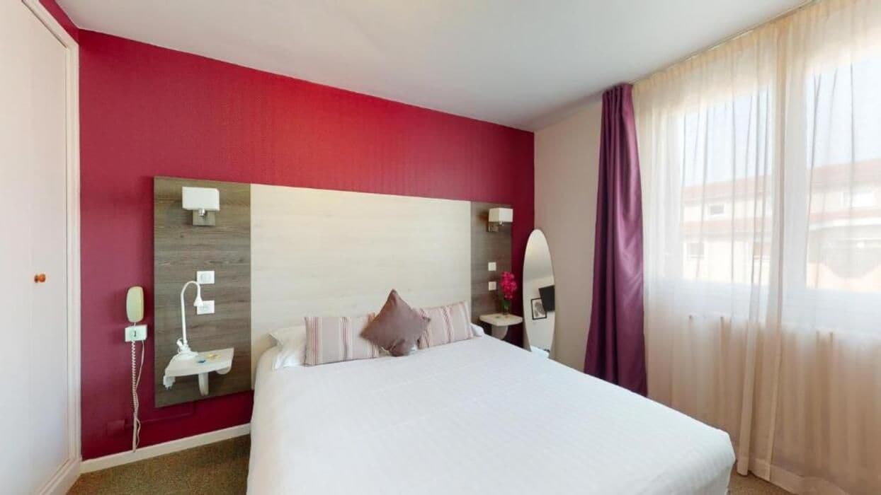 A hotel room with a large bed, a bedside table, a nightstand, a mirror, and a window with curtains.