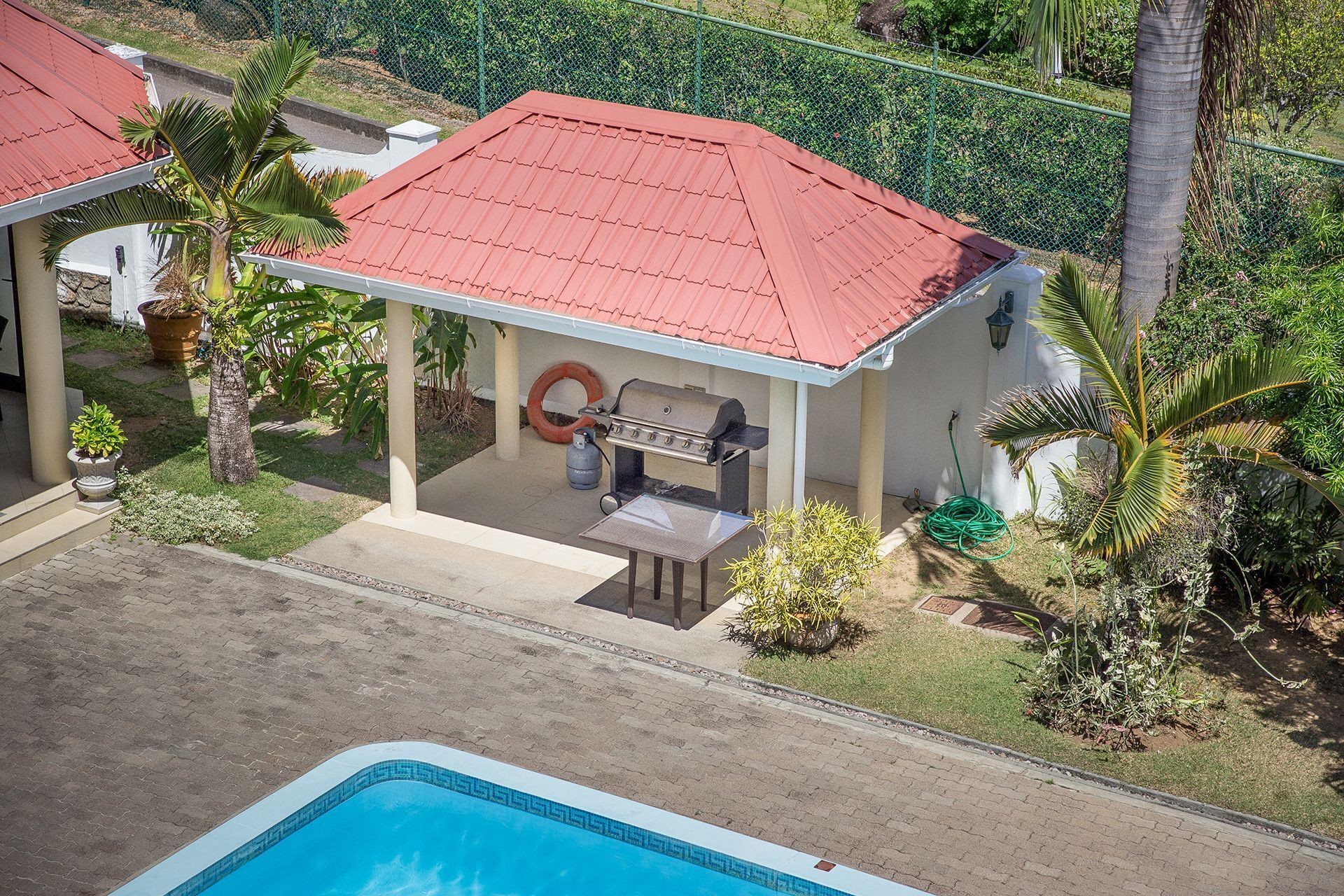 A covered outdoor area with a barbecue grill, a pool, and a red-roofed building.