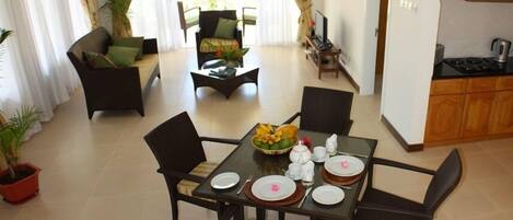 A dining area with a table set for four, surrounded by chairs. There is a sofa and a chair in the background, and a kitchen area is visible to the right.
