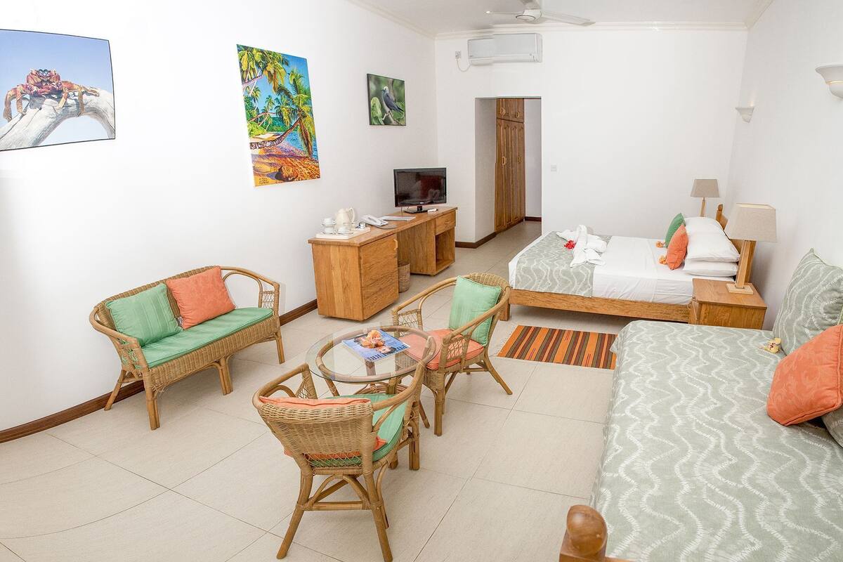 A room with a bed, two wicker chairs, a small table, a TV, and artwork on the walls.