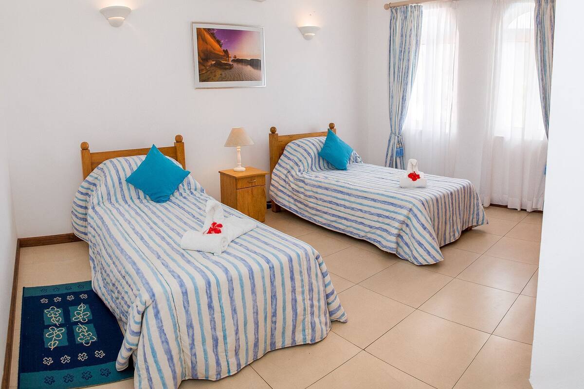 A hotel room with two beds, each with striped bedding and blue pillows, a nightstand with a lamp, a wall-mounted air conditioner, a framed picture, and a window with curtains.