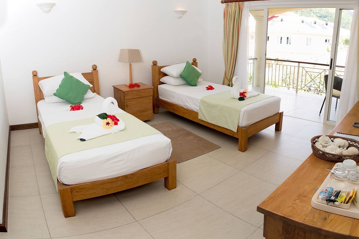 A hotel room with two wooden beds, each with a green pillow, white linens, and a towel with flowers. There is a nightstand with a lamp, a wooden table with breakfast items, and a view of the outside balcony.