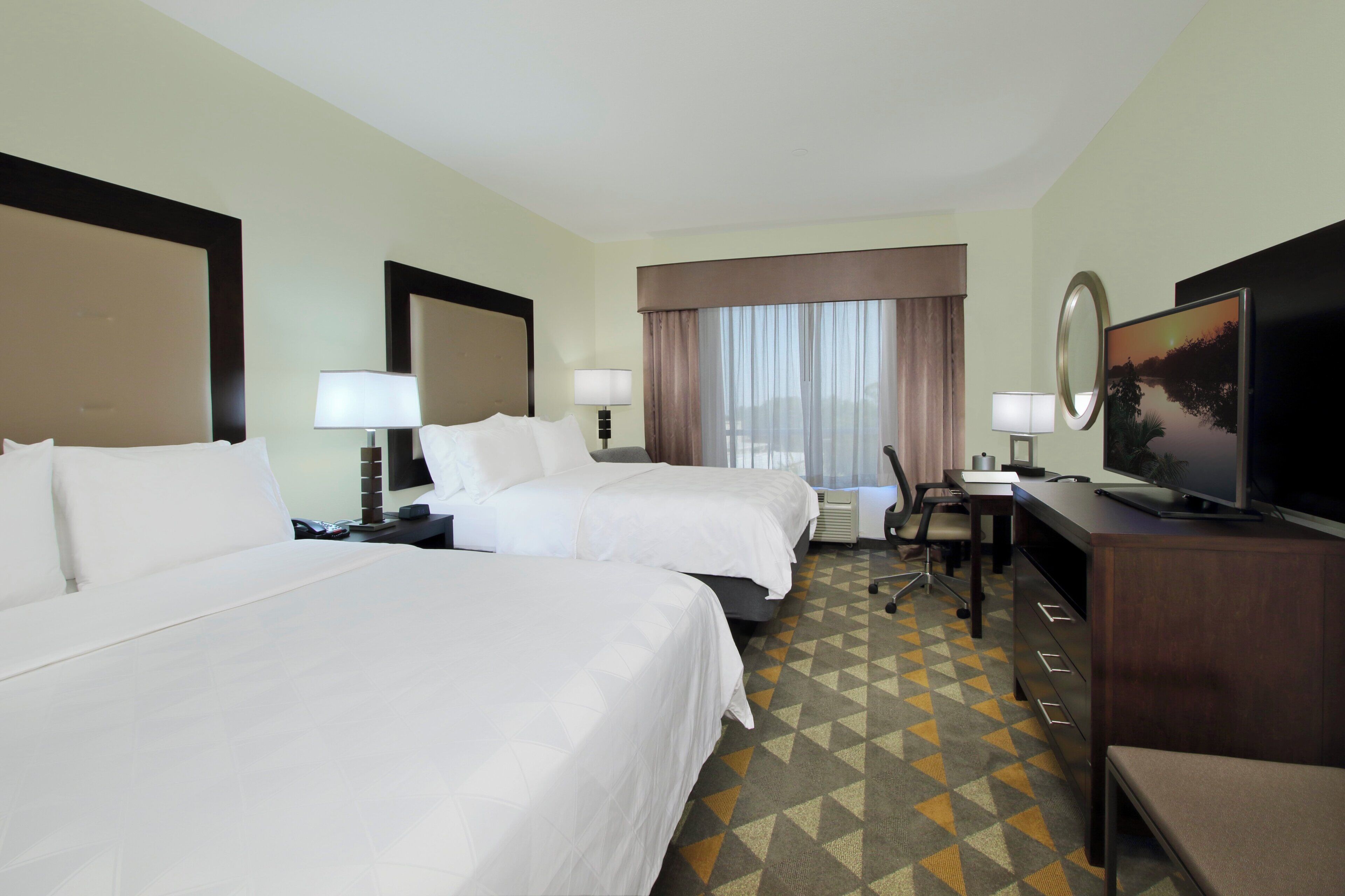 A hotel room with two beds, a desk, a chair, a television, and a large window with curtains.