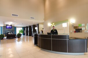 Zona de recepción de un hotel con mostrador, televisor de pantalla plana en la pared y un área de descanso con sofá y plantas en macetas.