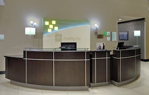 A reception desk with a glass partition, a flat-screen TV, and a digital display board.