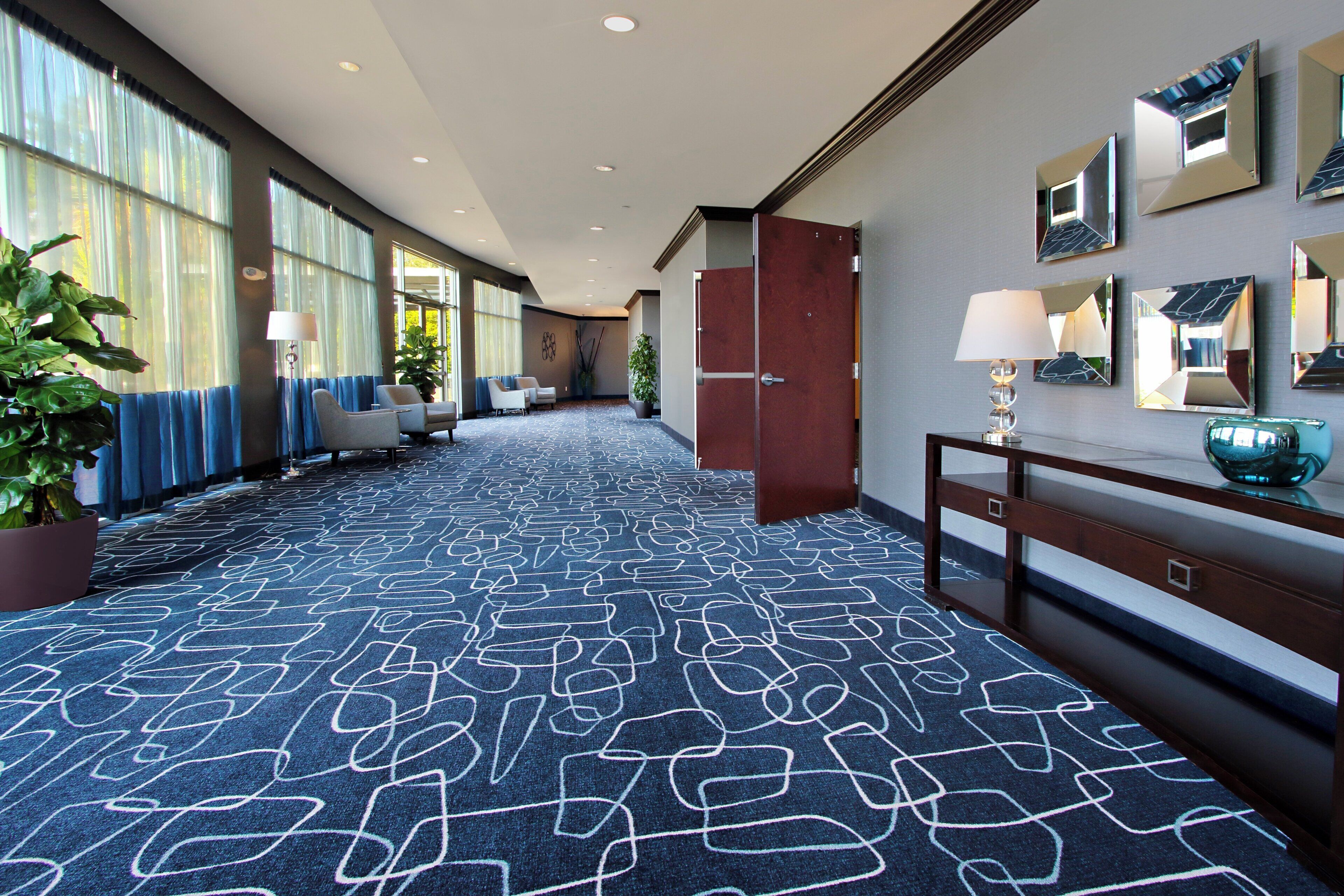 A hotel corridor with patterned carpet, seating area, potted plants, and wall-mounted mirrors.