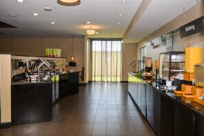 A modern hotel lobby with a bakery counter, a reception desk, and a large window with curtains.