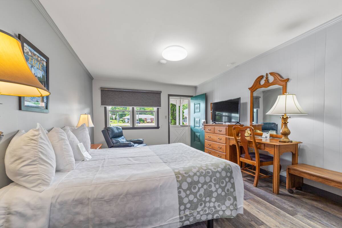 A bedroom with a bed, a desk, a chair, a TV, and a window.
