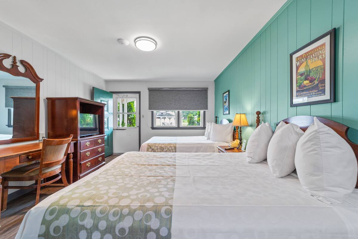 A hotel room with two beds, a desk, a chair, a television, and a window with a view of greenery.