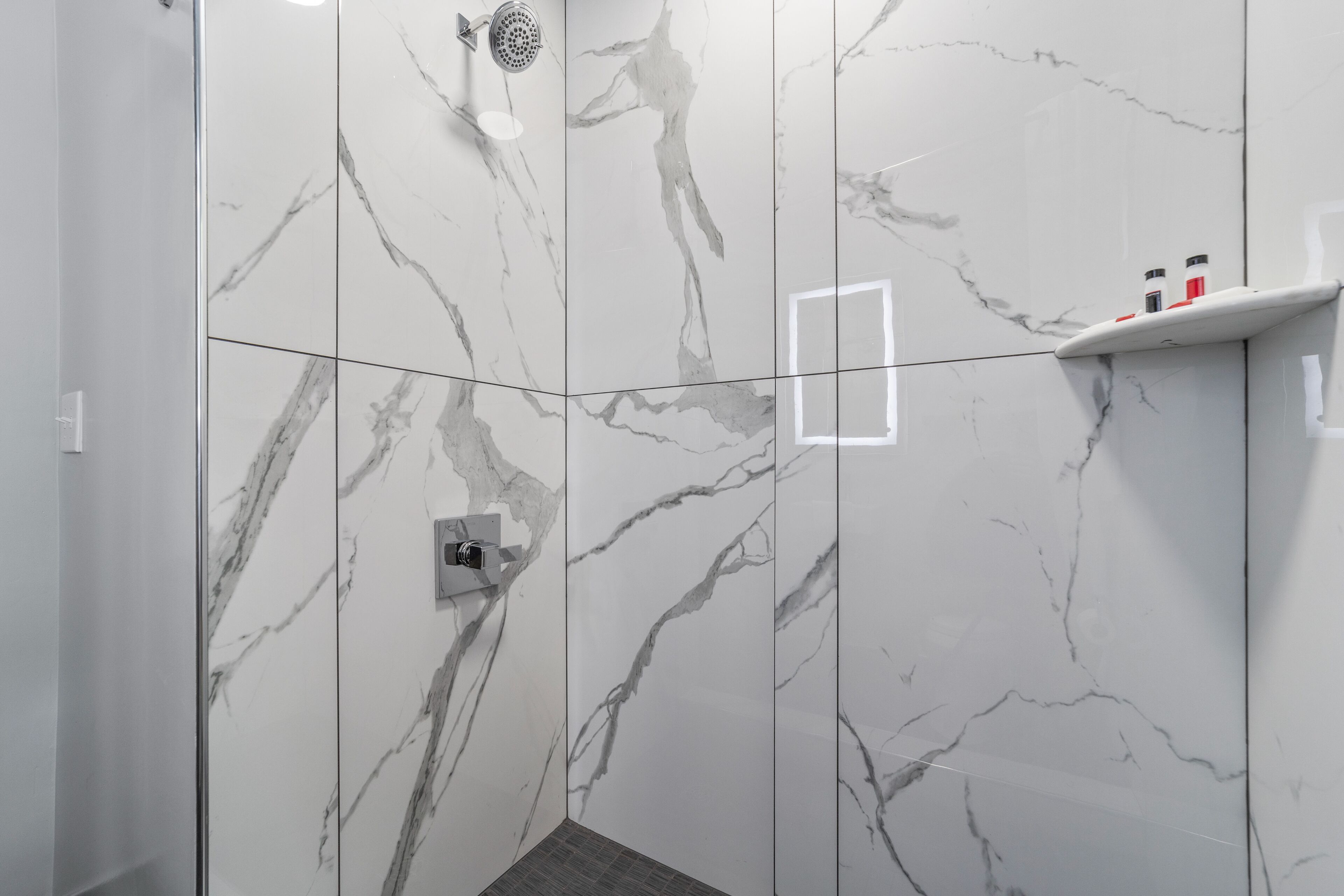 Baño con paredes de mármol, zona de ducha y repisa con artículos de aseo.